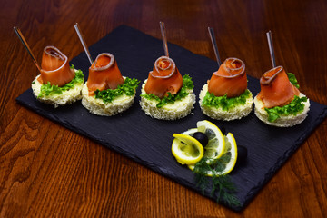 Cold appetizers, canape with red fish, wooden background. Canape with salad leaves, red fish, lemon and dill. Restaurant dish concept. Delicious snacks served in restaurant on black dishes.