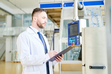 Profile view of concentrated bearded inspector wearing white coat taking necessary notes while carrying out inspection at dairy plant, portrait shot