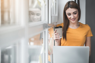 Portrait of girl using computers with pleasure. She sitting by the casement with gadgets on knees and holding beverage. Copy space in left side