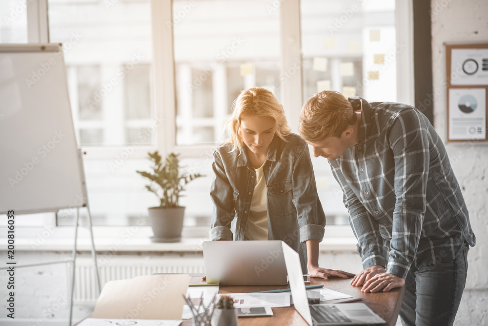 Poster man and woman standing in their working surrounding. both are staring at notebook monitor with seren