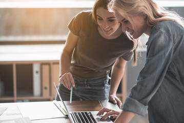 Young girls standing in office. Both are looking at notebook screen with delight