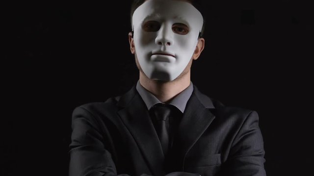 Anonymous suit man in mask folding hands on black background, illegal activity