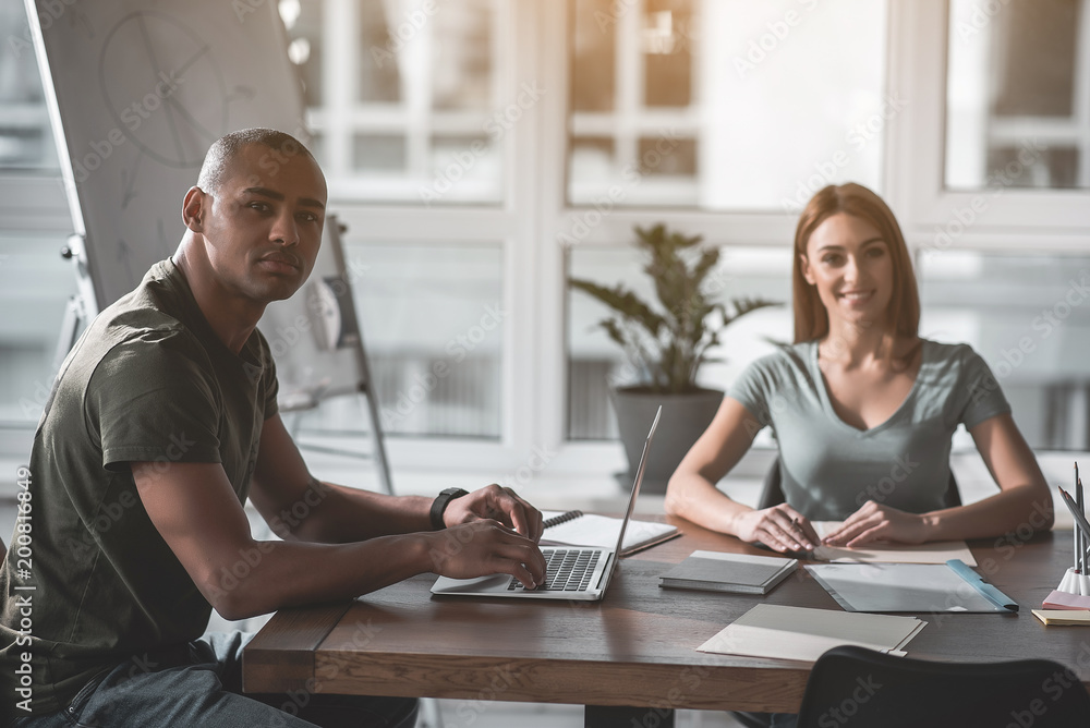 Poster portrait of pleased coworkers looking at camera while working at desk. guy holding hand on notebook 