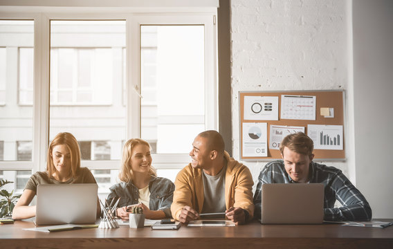 Men and women talking, using notebooks and smiling while sitting at desk. Window and board with graphics are on background