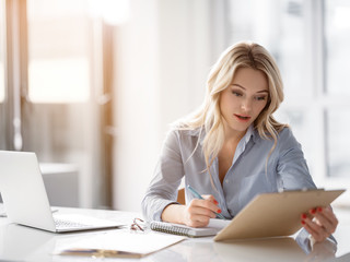 Portrait of serious young woman is reading document before signing it. She is sitting at desk in office