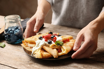 Woman putting plate with thin pancakes, berries and sour cream on table