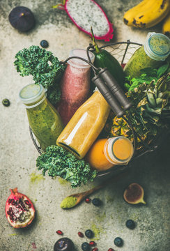 Colorful smoothies in bottles with fresh tropical fruit and greens in basket over grey concrete background, selective focus. Healthy, vegetarian, detox, clean eating breakfast food concept
