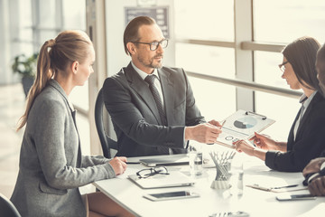 Calm male employer explaining graph while demonstrating it to comrades. They sitting at table indoor. Discussion during job concept