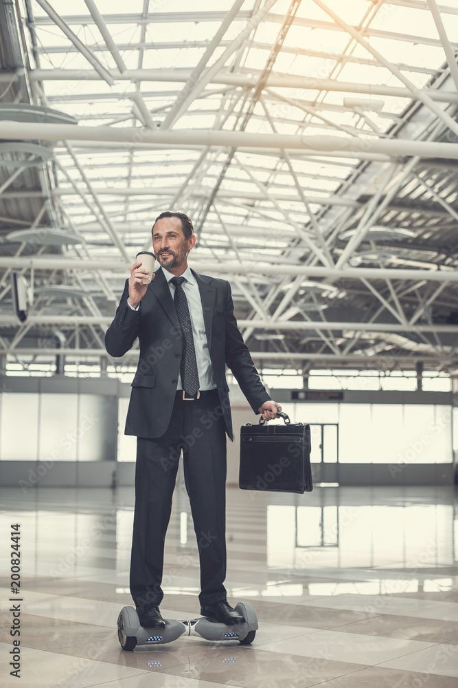 Poster full length portrait of cheerful businessman drinking mug of appetizing coffee while standing on gyr