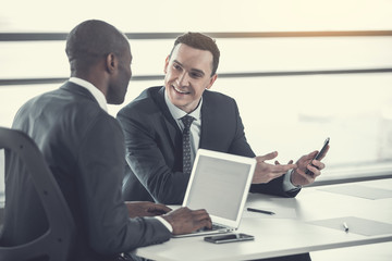 Fototapeta na wymiar Portrait of cheerful male telling with colleague while holding phone in arm. Employer typing in laptop. Beaming associates talking during job concept