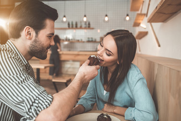 Affectionate young man is feeding his girlfriend with cake. Woman is biting with enjoyment and smiling