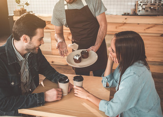 Your order, sir. Polite waiter is serving cakes and coffee to clients in cafe. Couple is sitting at table and smiling