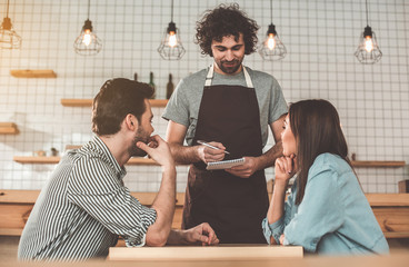 Great service. Portrait of joyful young waiter receiving an order from clients. He is writing into notebook and smiling