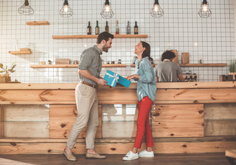 Full length of glad young man opening gift box in front of his girlfriend. Woman is standing with closed eyes and laughing. Barista is making coffee on background