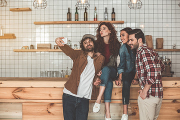 Portrait of joyful young men and women making selfie on cellphone. Guys are standing while girls are sitting on wooden table near them. Copy space