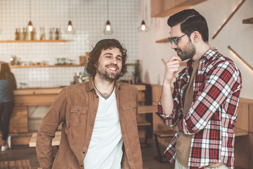 Portrait of happy young man telling story to his friend and laughing. Fellow is listening to guy attentively while standing in cafe indoors
