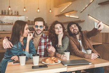 Happy young man is photographing with his friends on mobile phone. They are sitting at table in coffee shop and embracing