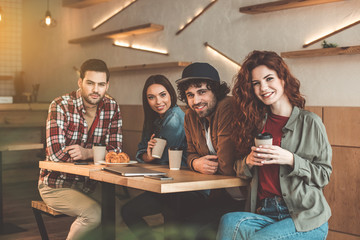Enjoying time with best friends. Portrait of happy young men and women drinking coffee in cafe. They are looking at camera and smiling