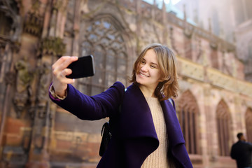 Young tourist making selfie mobile photo with famous Сathedral Notre Dame of Strasbourg on background - obrazy, fototapety, plakaty
