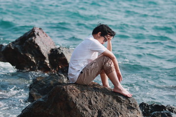 Upset depressed Asian man with hands on face feeling bad at rock of natural seashore