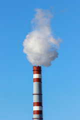 Thick white smoke from a high brick industrial plant chimney against a blue sky