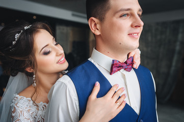A sweet bride straightens a bow tie to her fiancé. The bride takes care of the groom in the gray studio. Portrait of a newlywed couple.