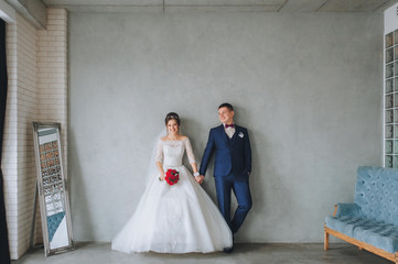 Cute newlyweds standing near the wall in a gray studio and holding hands cute smile. The bride and...
