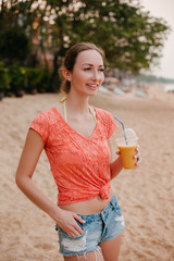 smiling attractive girl standing with cocktail on sandy beach and looking away