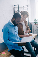 stylish businessman with paper cup of coffee watching on textbook and talking to african american coworker