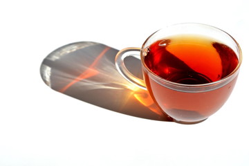 Hot black tea in transparent glass cup with long rainbow shadow in shaft of sunlight. Isolated. White background