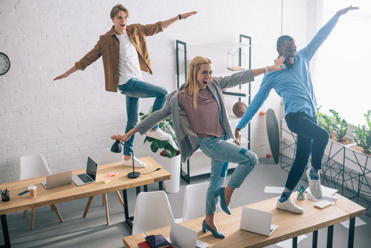 high angle view of happy multiethnic business colleagues dancing on tables in modern office