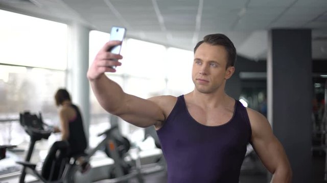 Bodybuilder taking selfie in gym and sending by smartphone, modern technology