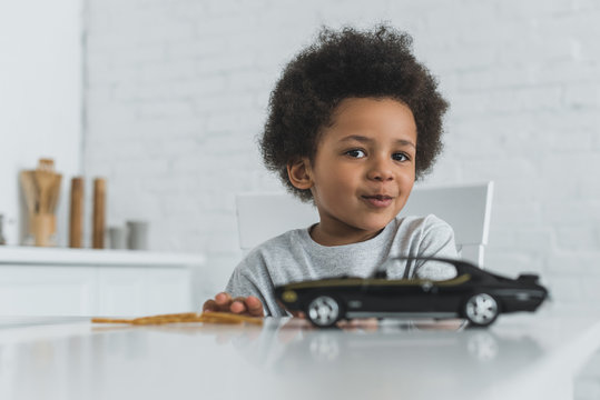 Adorable African American Boy Sitting At Table With Car Toy And Looking At Camera At Home