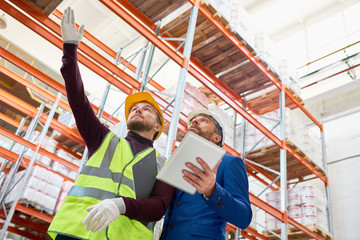 Low angle portrait of warehouse manager holding clipboard talking to worker wearing hardhat and reflective jacket while discussing stock inventory, pointing up