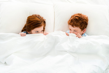 top view of cute little redhead children hiding under blanket and looking at each other