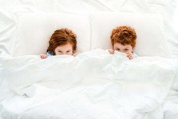 Obraz na płótnie Canvas top view of cute little redhead children hiding under blanket in bed