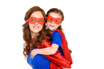 super mother piggybacking adorable daughter in mask and cloak and smiling at camera isolated on white