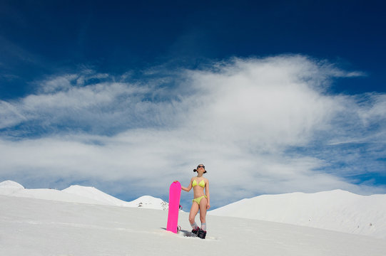 Sexy snowboarder woman in a swimsuit with a pink board standing on a mountain top