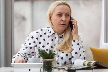 Beautiful adult woman with blond hair works in a cafe with a white interior. The company management by talking on the phone.