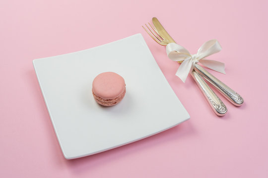 Silver fork and knife with white ribbon and plate with a maracon on pink background