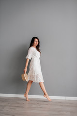 beautiful young girl in white short dress is holding in her hand a summer hat on a gray background. Fashion shooting