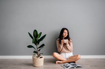 beautiful young brunette girl in white short shorts sitting on floor next to ficus on gray background in drinks coffee