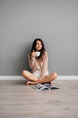 Fototapeta na wymiar beautiful young brunette girl in white short shorts sitting on floor on gray background and drinking coffee
