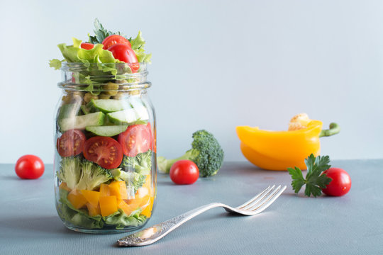 Vegetable healthy homemade colorful salad in mason jar with tomato, lettuce, broccoli on blue. Copy space. Lunch for work. Food set.