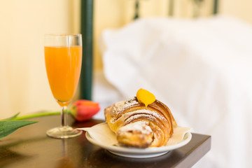 Sweet breakfast in hotel room. Croissant with yellow cream, glass of orange juice and flower on the night table 