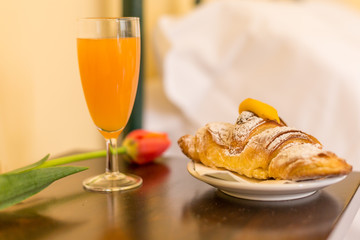 Close-up of a croissant with yellow cream and icing sugar, glass of orange juice and red flower on the night table. Sweet breakfast