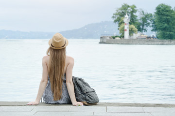 Girl in the hat sitting on the dock. Sea and small lighthouse in the distance. Back view