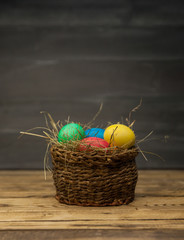 Easter eggs of different colors in a wicker basket with hay on a wooden background
