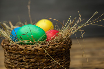 Easter eggs of different colors in a wicker basket with hay on a wooden background