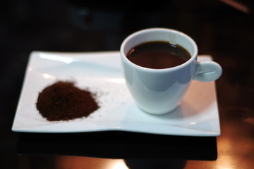 Hot coffee on white ceramic glass and fresh roasted coffee beans on a white plate on a brown table.